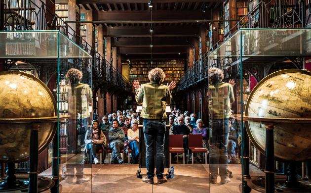Sfeerbeeld Nottebohmlezing Dirk Draulans aan spreekgestoelte, globes in voorgrond, publiek in achtergrond